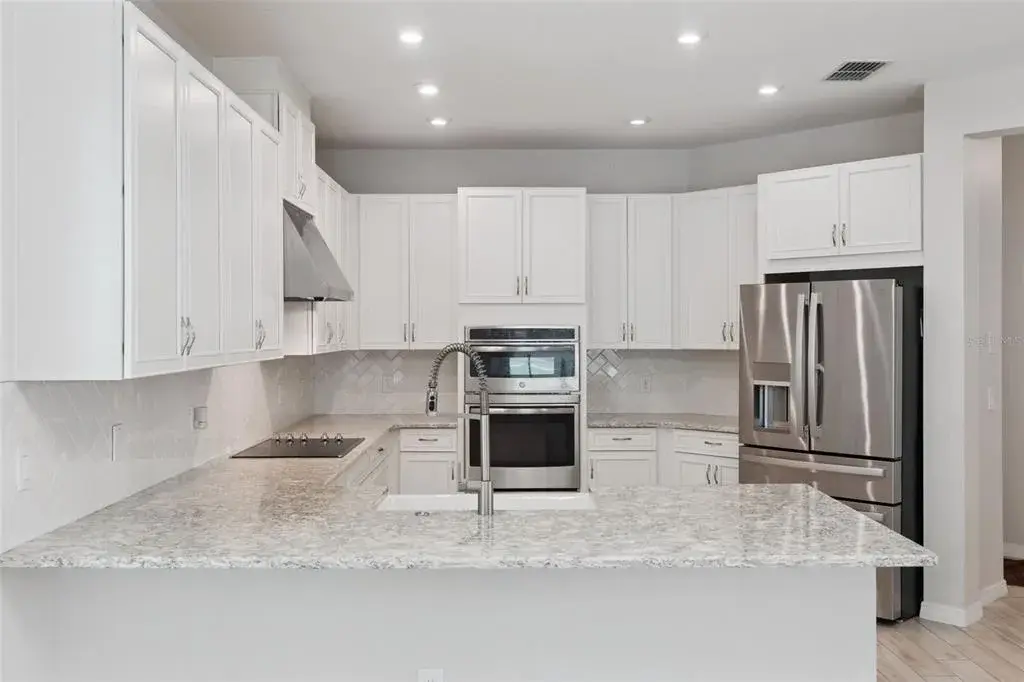 Kitchen – Spacious kitchen features abundant white cabinetry, granite countertops, a classic tile backsplash, stainless steel appliances, double wall ovens, and a generous peninsula with farmhouse-style sink and statement faucet.
