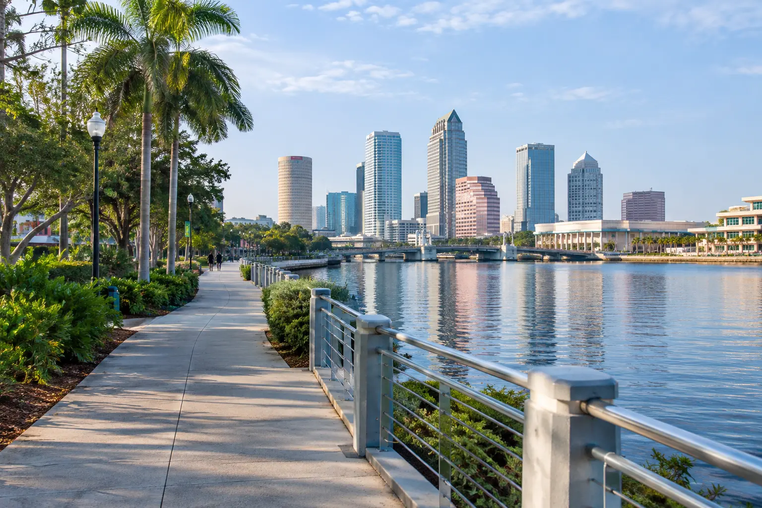 Morning view along the Tampa Riverwalk, with pedestrians, waterfront greenery, the river and downtown Tampa in the background.