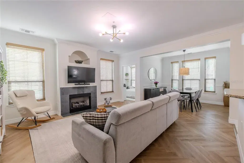 The living room has a bright, open layout with wide-plank flooring laid in a subtle herringbone pattern adding warmth and visual interest, while crisply painted walls and crown molding create a clean, timeless backdrop. Natural light filters in through multiple windows fitted with wood-look blinds, giving the space an airy, well-balanced feel throughout the day.