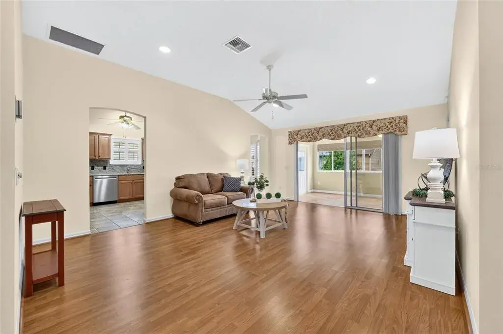 Bright, spacious living room featuring vaulted ceilings, wood-look flooring, recessed lighting, and sliding glass doors leading to the enclosed lanai.