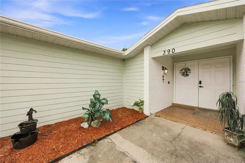 Welcoming covered front entry featuring double white front doors, tiled threshold, and mulch landscaping bed with water feature.