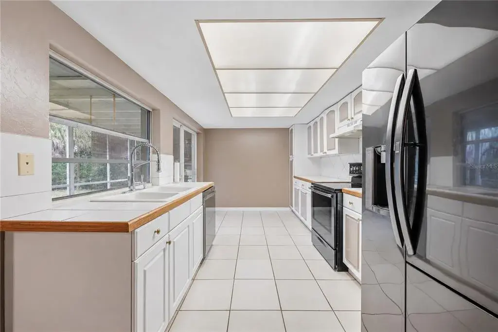 Open-concept kitchen highlighting white cabinetry, stainless steel refrigerator, tile backsplash, and pass-through window to the lanai.