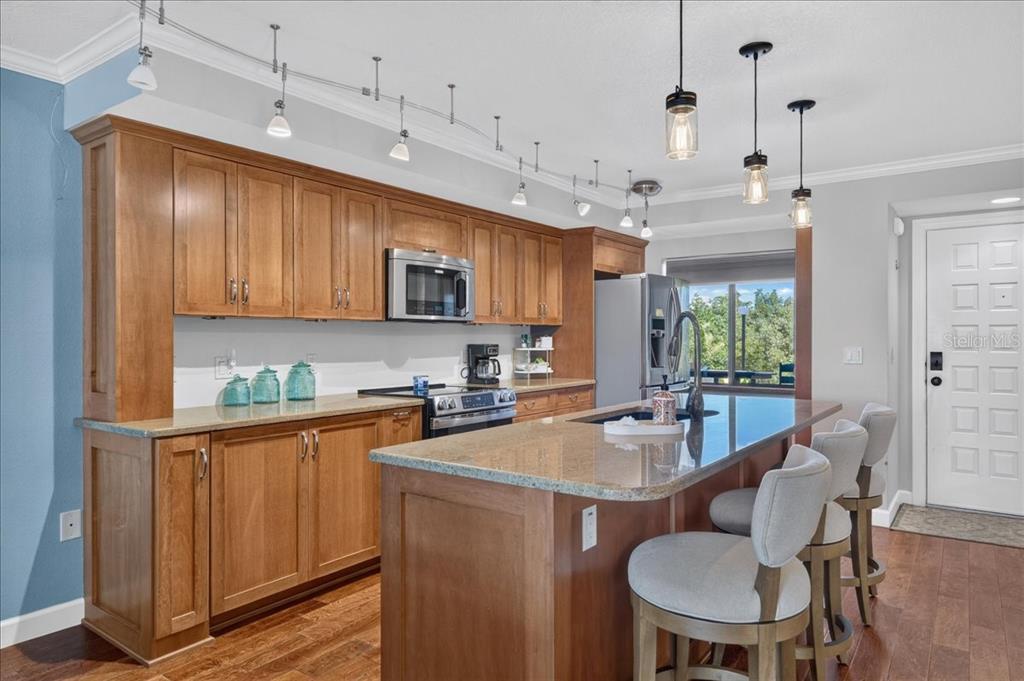 The kitchen features custom solid wood soft-close cabinetry, granite countertops, high-end appliances, and a large island with seating, all flowing naturally into the dining and living areas.