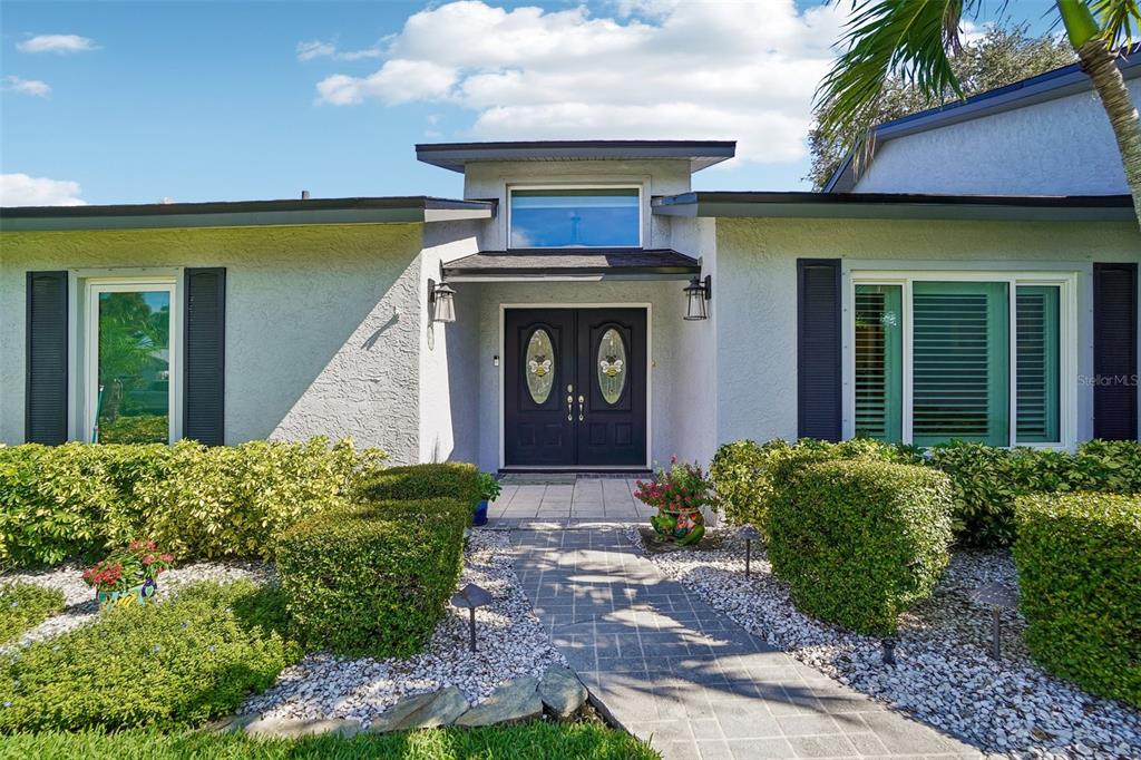 Front Entry – A paved walkway surrounded by manicured landscaping leads to decorative double entry doors, accented by exterior lighting and a large window above.