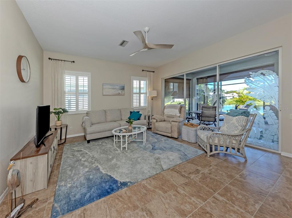 A spacious, inviting living room featuring beautiful, seamless ceramic tile flooring and elegant plantation shutters throughout the home, along with stunning etched-glass sliding doors opening to the extended lanai, bringing the beauty of the outdoors right into view!