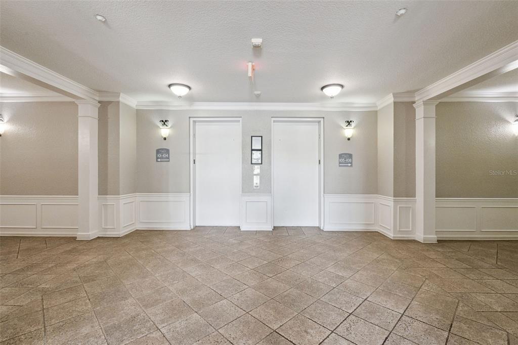 Elevator Lobby:Spacious interior building lobby featuring tiled flooring, classic wainscoting, warm wall sconces, and dual elevator access.