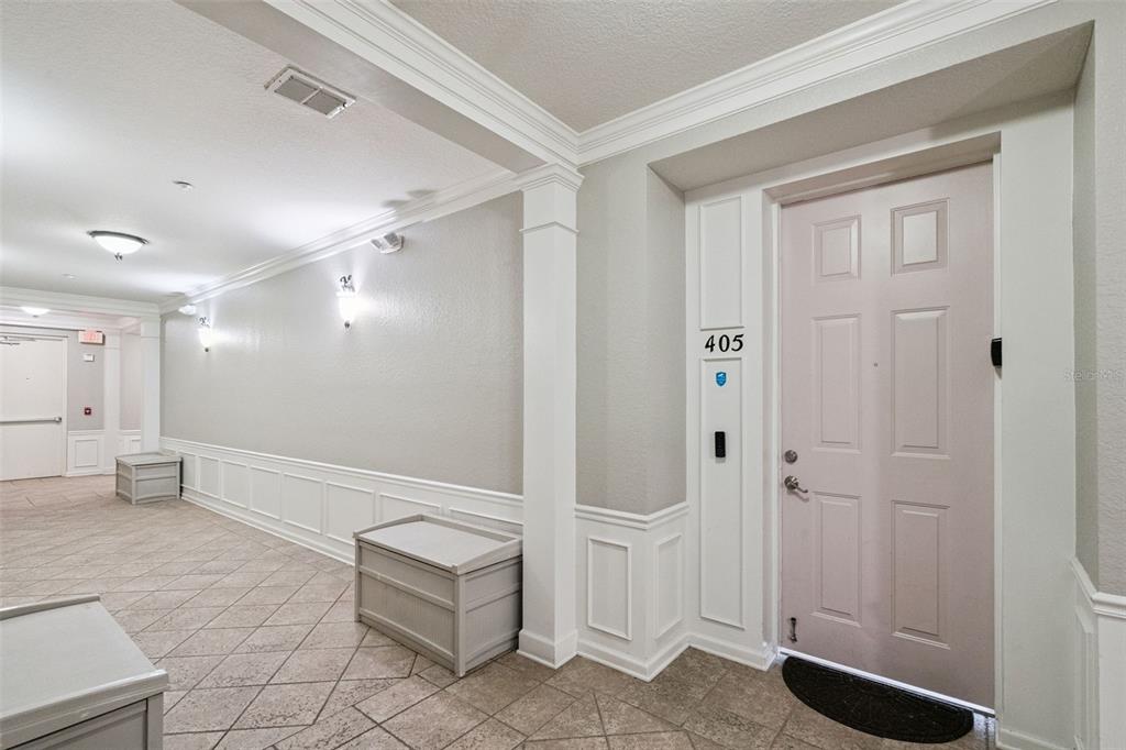 Interior Hallway:Well-lit interior corridor showcasing tiled flooring, wainscoting trim, storage benches, and private entrance to Unit 405.