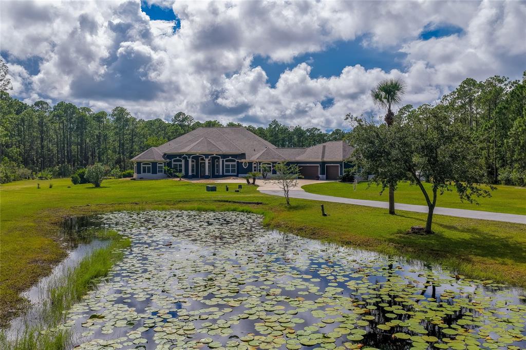 Foreground view of a lily-pad-covered pond looking toward a sprawling home framed by a paved driveway and mature pine trees.