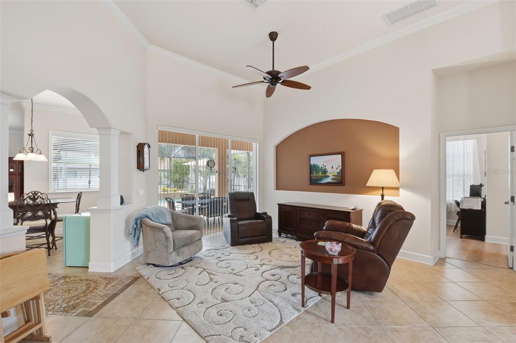 Living Room – Spacious living area with high ceilings, crown molding, a ceiling fan, and architectural archways that create an open connection to adjoining spaces. Sliding glass doors bring in natural light and provide access to the outdoor Lanai and Pool.