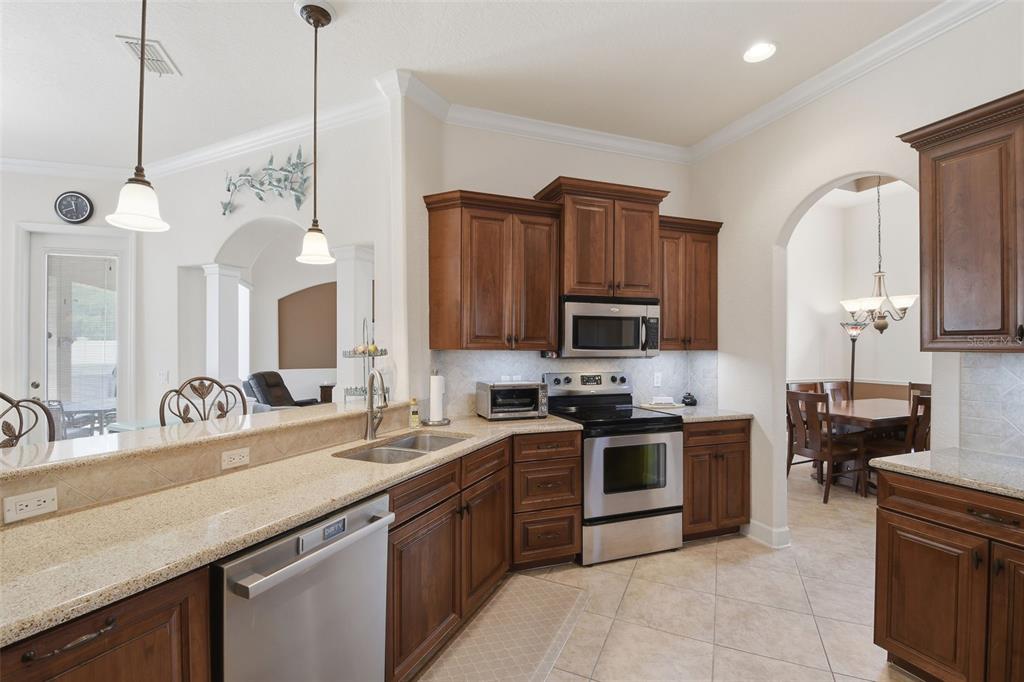 Kitchen – Well-appointed kitchen featuring warm wood cabinetry, granite countertops, stainless steel appliances, tile backsplash, and a breakfast bar that opens toward the adjacent living space.